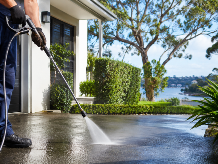 A PeakWash exterior cleaning technician pressure cleaning a residential driveway in Gymea Bay NSW, with water spraying across the surface beside a clean home, trimmed hedges, native trees and bay views in the background.