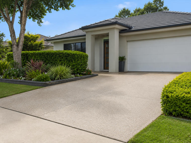 Front view of a freshly cleaned residential driveway in Sylvania NSW, showing a tidy modern home, clean garage, landscaped garden beds, green hedges and neat lawn under a bright blue sky.